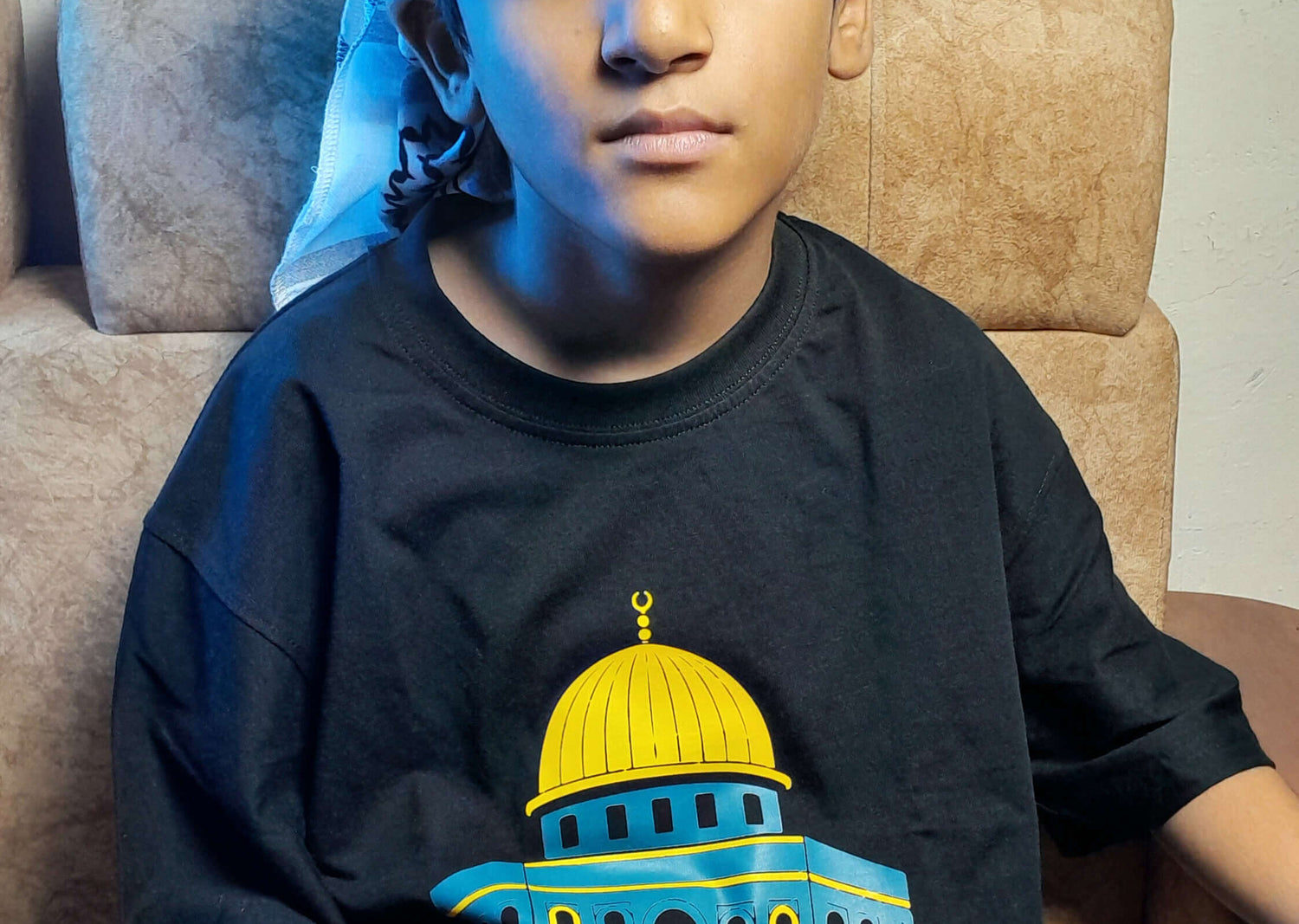 Child wearing a black t-shirt with a Dome of the Rock design, sitting on a beige chair.